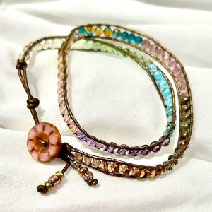 Könnte beinhalten: Ein mehrreihiges Armband mit einem rosa Knopfverschluss. Das Armband verfügt über eine Vielzahl von bunten Perlen, darunter lila, blau, grün und braun.