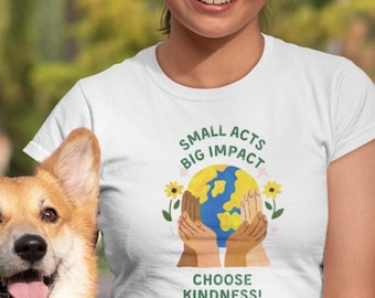 May include: A woman wearing a white t-shirt with a graphic of a globe surrounded by hands and the text "Small Acts Big Impact Choose Kindness!" sitting on the grass with a corgi dog.