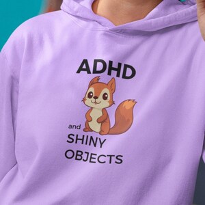 Puede incluir: Una sudadera con capucha lavanda con una ardilla de dibujos animados y el texto "TDAH y OBJETOS BRILLANTES".