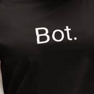 May include: A black long-sleeve shirt featuring the word Bot. printed in white capital and lowercase letters across the chest. The garment has a crew neckline and long sleeves, providing a simple and clean design for casual everyday wear.
