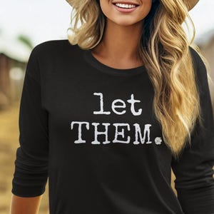 May include: Black long-sleeve shirt with the text "let THEM." in white. The shirt is paired with a brown belt with a decorative buckle and light-colored jeans. A beige cowboy hat completes the look.