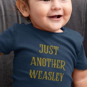 May include: Navy blue baby bodysuit with the text "JUST ANOTHER WEASLEY" in gold lettering. The short-sleeved onesie is worn by a smiling baby sitting on a gray couch.