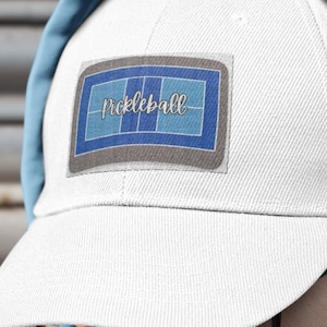 May include: A white baseball cap with a blue and grey patch that says "pickleball" on it. The cap is being worn by a person wearing a light blue hoodie.