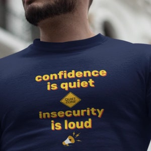 以下が含まれることがあります： 「confidence is quiet」というテキストと黄色の「Quiet Zone」サインが書かれたネイビーブルーのTシャツ。サインの下には「insecurity is loud」というテキストと黄色のメガホンが描かれています。