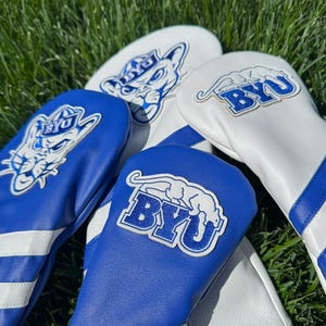 May include: Golf club head covers in blue and white, featuring the BYU logo. The covers are made of a leather-like material and are arranged on a bed of green grass. The covers have the BYU logo and stripes.