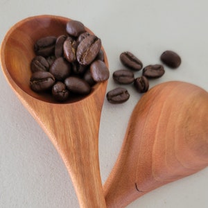 Könnte beinhalten: Zwei Holzlöffel mit einem Loch im Griff. Ein Löffel ist mit Kaffeebohnen gefüllt, und ein paar Kaffeebohnen sind auf der weißen Oberfläche verstreut.