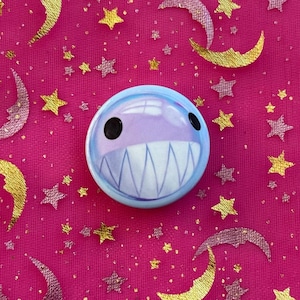 May include: A round, light blue button with a cartoon shark face, featuring black eyes and a wide, toothy grin. The button is set against a vibrant pink background adorned with gold and silver crescent moons and stars.