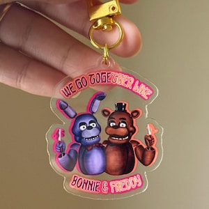May include: A clear acrylic keychain featuring Bonnie and Freddy characters from the Five Nights at Freddy's franchise. The keychain has a gold-colored clasp and the text "WE GO TOGETHER LIKE BONNIE & FREDDY" in pink.
