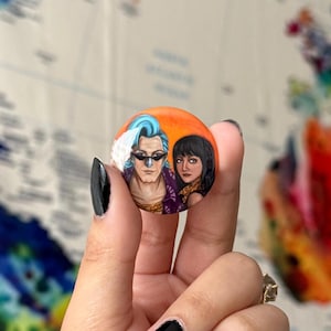 May include: A round button with a painted image of two people. The man has blue hair and is wearing sunglasses. The woman has long dark hair and is wearing a purple and black outfit. The background is orange.
