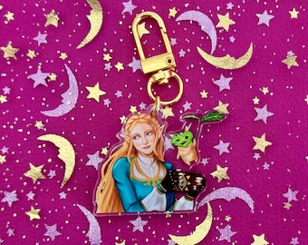 May include: A clear acrylic keychain featuring a character from a video game, with long blonde hair and a blue and gold outfit. The keychain has a gold-colored clasp and a small green and brown creature charm. The background is a pink fabric with gold and silver stars and moons.