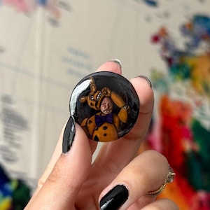 May include: A black button with a round image of a person wearing a brown and yellow animatronic costume. The person is smiling and holding the animatronic head in front of their face.