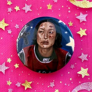 May include: A round button pin featuring a portrait of a person with blood splatters on their face. The background of the pin is a blue night sky with white stars. The person is wearing a red and white top. The background is pink with gold stars.