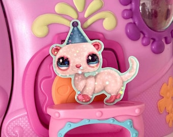May include: A pink toy ferret figurine with a blue party hat and star accents. The figurine is standing on a pink platform with a blue trim, set against a backdrop of a pink toy playset with decorative elements.