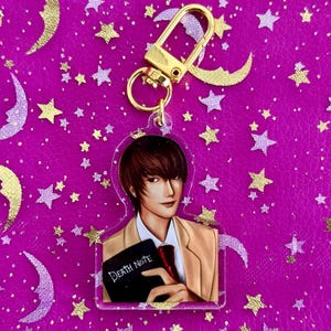 May include: Acrylic keychain featuring an anime character holding a black notebook with "Death Note" text. The character wears a tan blazer and red tie. The keychain has a gold-colored clasp and is set against a pink background with star and moon shapes.