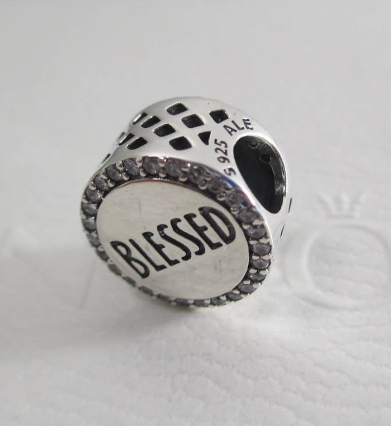 Puede incluir: Encanto de plata con un borde de cristal transparente y la palabra "Blessed" en letras negras. El encanto tiene un patr&oacute;n de celos&iacute;a decorativo en la parte posterior y la inscripci&oacute;n "S 925 ALE" en el lateral.