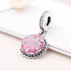 May include: A silver charm with a pink cake design that says "Happy Birthday". The charm has a silver loop at the top for attaching to a bracelet or necklace.