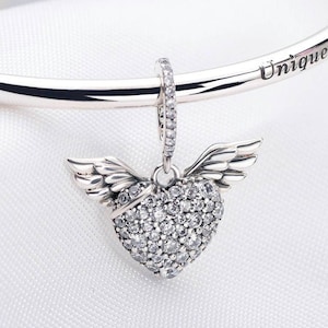 May include: A silver charm with a heart-shaped pendant covered in clear cubic zirconia stones. The heart has wings on either side and is attached to a silver chain.