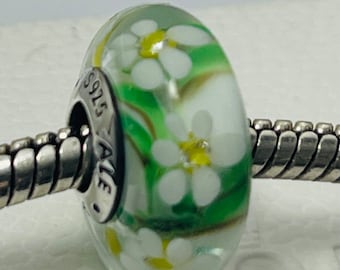 May include: A silver charm bead with a green and white glass charm featuring a white daisy pattern. The charm is marked with the text "925 ALE".