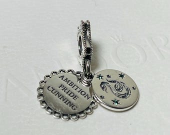 May include: Silver charm with two circular pendants. One pendant features a snake with green gemstones and stars. The other pendant has a scalloped edge and the words "Ambition, Pride, Cunning".