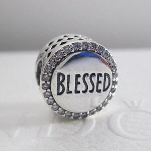 Puede incluir: Encanto de plata con un borde de cristal transparente y la palabra "BLESSED" en letras negras.