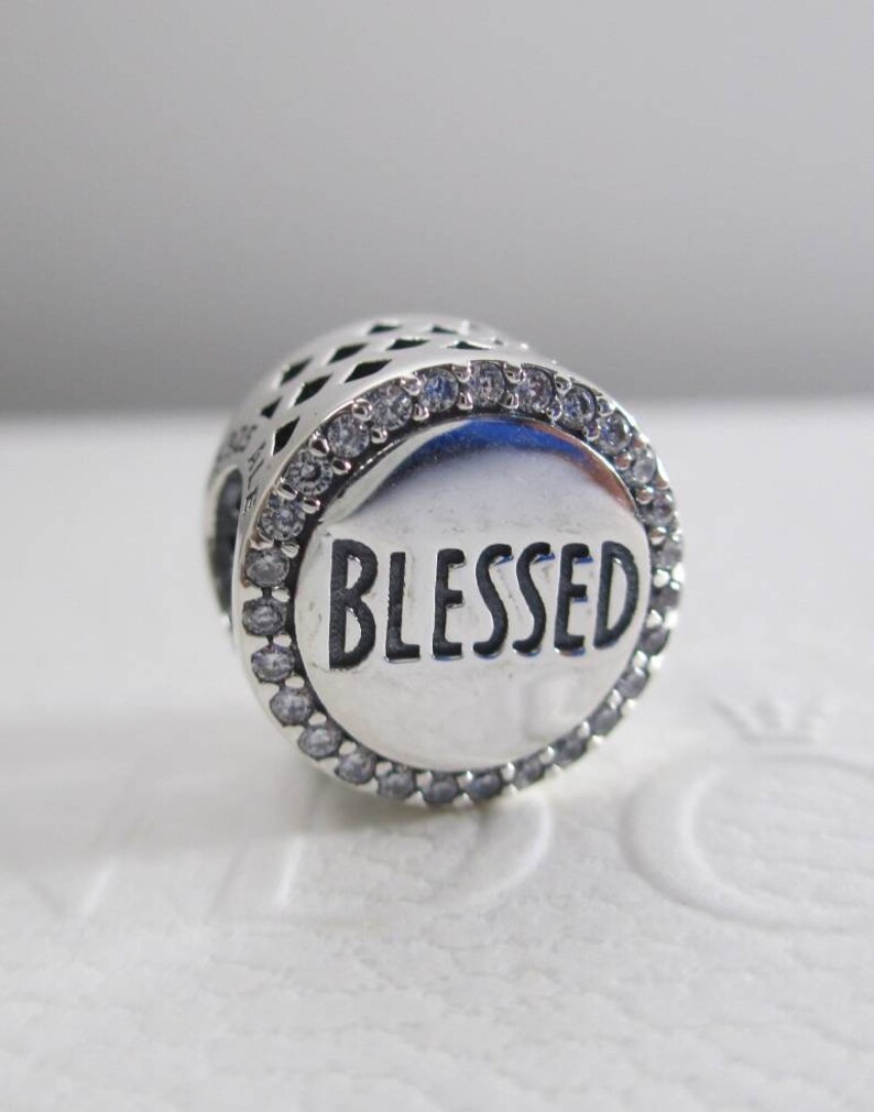Puede incluir: Encanto de plata con un borde de cristal transparente y la palabra "BLESSED" en letras negras.