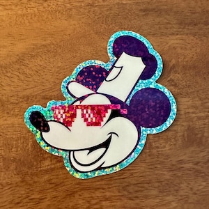 May include: A glittery sticker of Mickey Mouse wearing red and white pixelated sunglasses. The sticker has a blue and purple iridescent finish.