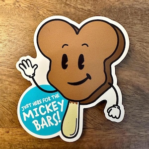 May include: A brown cartoon ice cream bar with a smiling face and stick, waving with a white gloved hand. The ice cream bar is on a blue circle with the text "Just here for the Mickey Bars!"