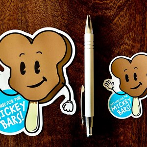 May include: Two brown and white cartoon ice cream bars with a smiling face and arms. The larger ice cream bar has a blue circle with the text "Just here for the Mickey Bars!" The smaller ice cream bar has a blue circle with the text "Just here for the Mickey Bars!"