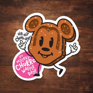 Peut inclure: Une gaufre en forme de Mickey Mouse de dessin animé avec de la sauce au chocolat et des pépites. La gaufre porte un gant blanc et a une bulle de dialogue rose qui dit "Hip, Hip Churr-ay! It's Mickey Churro Waffle Day!"