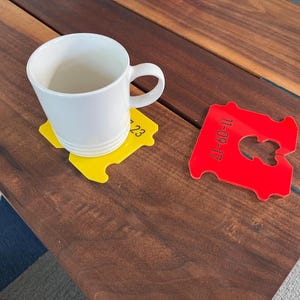 May include: A white ceramic mug rests on a yellow coaster with the number "23" printed on it. A red bread tag coaster with the date "11-09-17" is also present. The coasters are on a dark wooden table.
