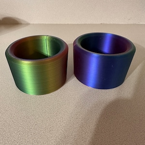 May include: Two iridescent 3D printed cups, one green and one purple, on a beige surface.