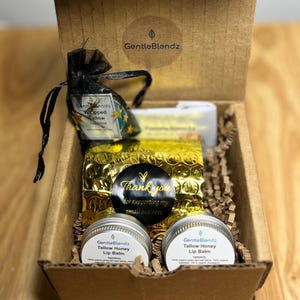 May include: A brown cardboard gift box containing two round silver tins of GentleBlendz Tallow Honey Lip Balm, a black organza bag labeled Whipped Tallow Jasmine, and a gold bubble wrap pouch with a black sticker that reads Thank you for supporting my small business.