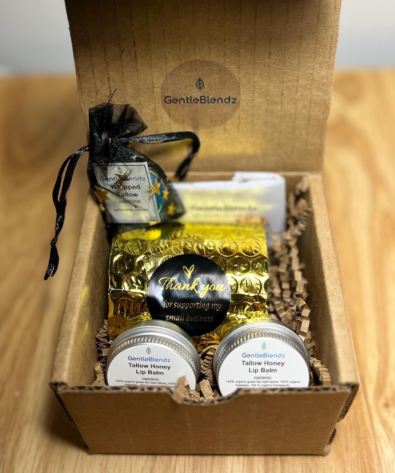 May include: A brown cardboard gift box containing two round silver tins of GentleBlendz Tallow Honey Lip Balm, a black organza bag labeled Whipped Tallow Jasmine, and a gold bubble wrap pouch with a black sticker that reads Thank you for supporting my small business.