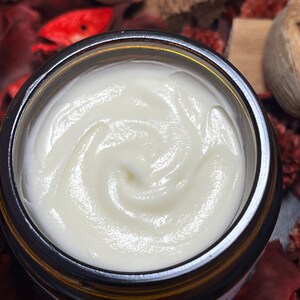 May include: A top-down view of an open amber glass jar filled with a thick, smooth, white cream. The cream has a swirled texture on the surface. The jar features a partial label with visible text, and the container is surrounded by dried botanicals.