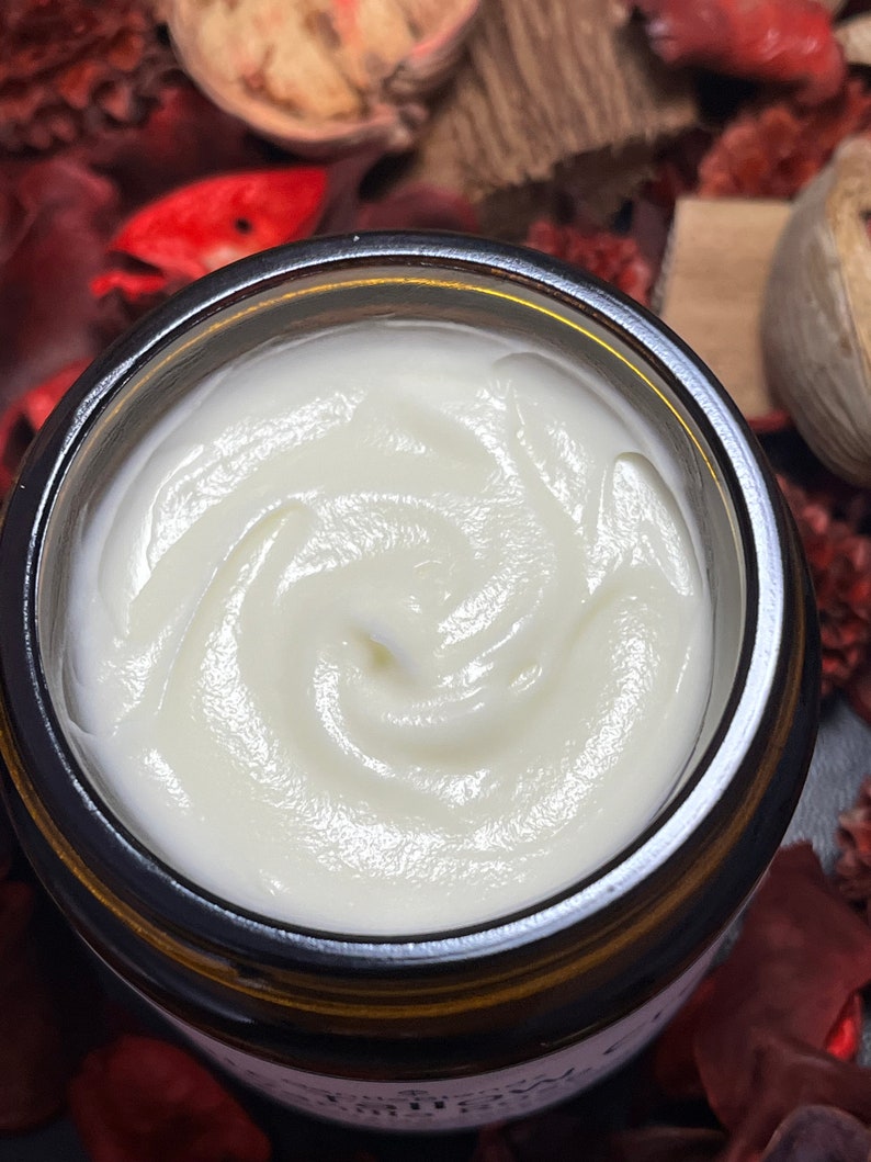 May include: A top-down view of an open amber glass jar filled with a thick, smooth, white cream. The cream has a swirled texture on the surface. The jar features a partial label with visible text, and the container is surrounded by dried botanicals.