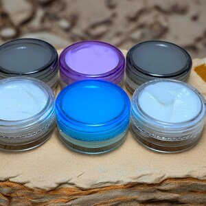 May include: Six small, clear jars with colorful lids and contents. The lids are gray, purple, and blue. The contents appear to be creams or gels in shades of white, light blue, and purple. The jars are arranged on a textured, tan surface.