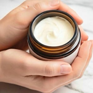 May include: A small, round, amber-colored glass jar with a white label, filled with a thick, smooth, off-white cream. The jar has a dark brown rim and is open at the top, revealing the textured, swirled surface of the cream inside the container.