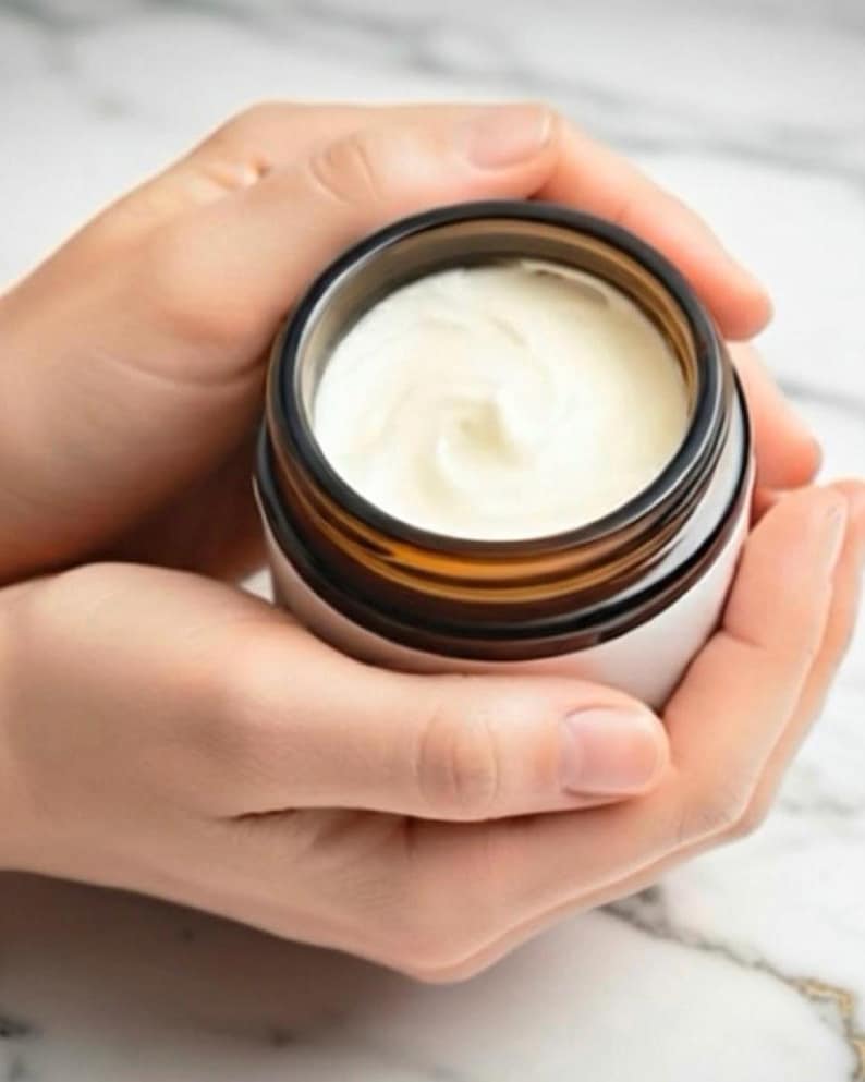 May include: A small, round, amber-colored glass jar with a white label, filled with a thick, smooth, off-white cream. The jar has a dark brown rim and is open at the top, revealing the textured, swirled surface of the cream inside the container.