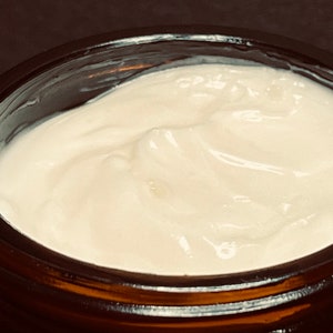 May include: A thick, white, opaque cream with a smooth, slightly swirled texture contained within a dark amber glass jar. The cream fills the circular opening of the jar, displaying a consistent, dense, and creamy appearance throughout the surface.