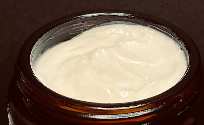 May include: A thick, white, opaque cream with a smooth, slightly swirled texture contained within a dark amber glass jar. The cream fills the circular opening of the jar, displaying a consistent, dense, and creamy appearance throughout the surface.