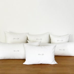 Op de afbeelding: Zes witte kussenhoezen in verschillende maten. De kussenhoezen zijn in een rij op een houten oppervlak gerangschikt. De maten van de kussens zijn aangegeven in inches: 24 x 24, 22 x 22, 20 x 20, 14 x 42, 14 x 36 en 14 x 20.