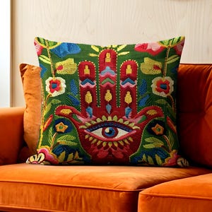 May include: Embroidered decorative pillow with a Hamsa hand design. The pillow features a green background with a red hand, an eye, and colorful floral accents in blue, yellow, and pink. The pillow is square and rests on an orange velvet sofa.