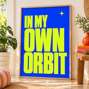 Könnte beinhalten: Ein rechteckiges Wandbild in einem hellen Holzrahmen mit einem leuchtend blauen Hintergrund und fett gedrucktem, hellgelbem Text, der IN MY OWN ORBIT lautet, ergänzt durch ein kleines, vierzackiges gelbes Sternsymbol in der oberen rechten Ecke.