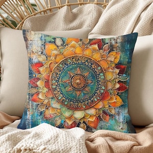 May include: Square decorative pillow with a colorful mandala design. The pillow features a central teal circle with a starburst pattern, surrounded by layers of orange, yellow, and red petal-like shapes. The background has a textured, distressed look with blue and teal hues.