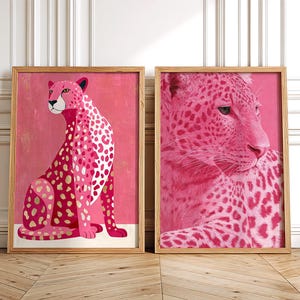 May include: Two framed art prints featuring pink cheetahs. One print shows a full cheetah, the other a close-up of its face. The cheetahs have white and pink spots with gold accents, set against a pink background.
