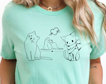 May include: A mint green t-shirt with a graphic of three cats. The cats are drawn in black line art and one cat is wearing a scarf. The graphic includes a heart above the cats.
