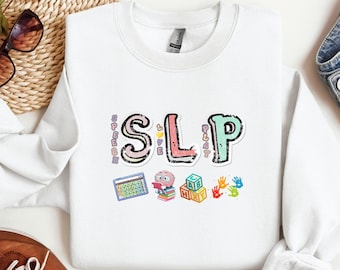 May include: White sweatshirt with a colorful graphic that says "SLP" with the words "Speech, Language, Play" written around the letters. The graphic also includes a book, a calculator, blocks, and handprints.