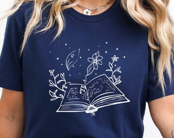 Puede incluir: Una camiseta azul marino con un gráfico blanco de un libro abierto con flores y hojas. El texto "Navy" está en la esquina superior izquierda de la imagen.