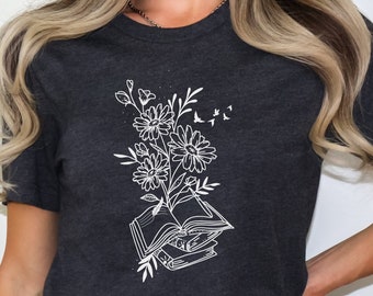 Puede incluir: Una camiseta de color gris oscuro jaspeado con un diseño gráfico blanco de una pila de libros con flores que crecen en la parte superior. Los pájaros vuelan por encima de las flores.