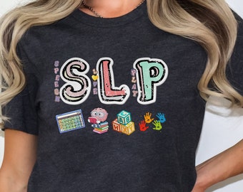 Op de afbeelding: Donkergrijs heather t-shirt met het woord "SLP" in kleurrijke letters. De letters worden omringd door afbeeldingen die te maken hebben met logopedie, waaronder een spraakballon, een boek, blokken en handafdrukken.